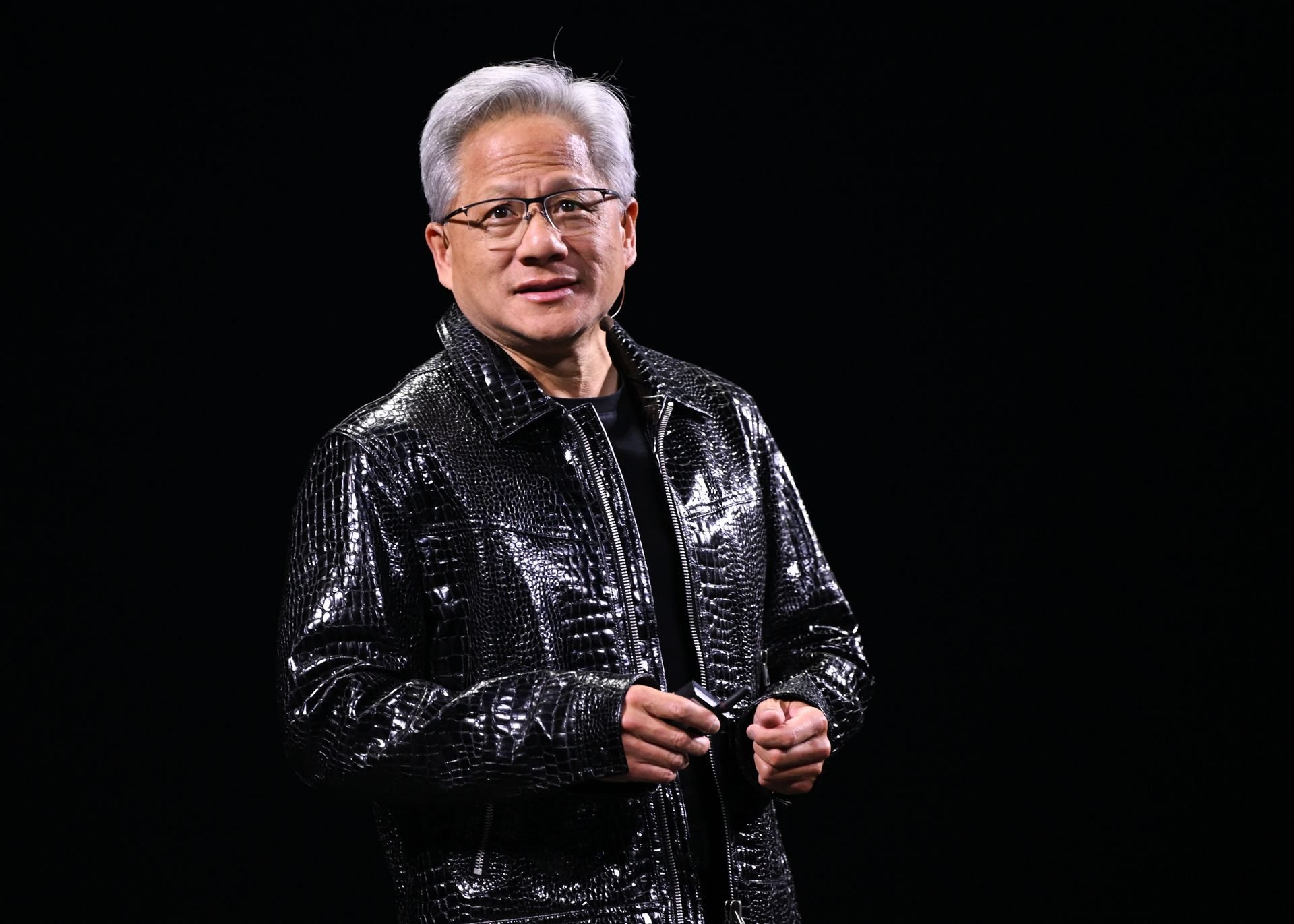 a close-up photo of Nvidia CEO Jensen Huang standing on stage in a black leather jacket in front of a black screen