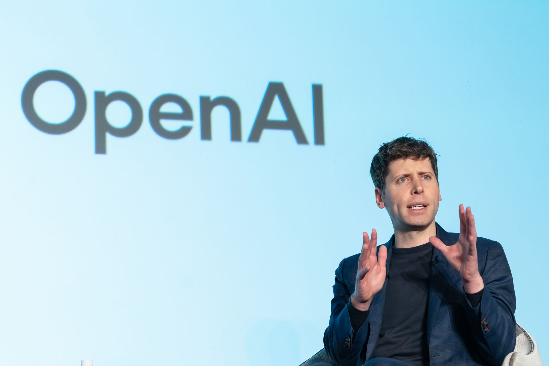Sam Altman sits in front of a screen that reads "OpenAI"