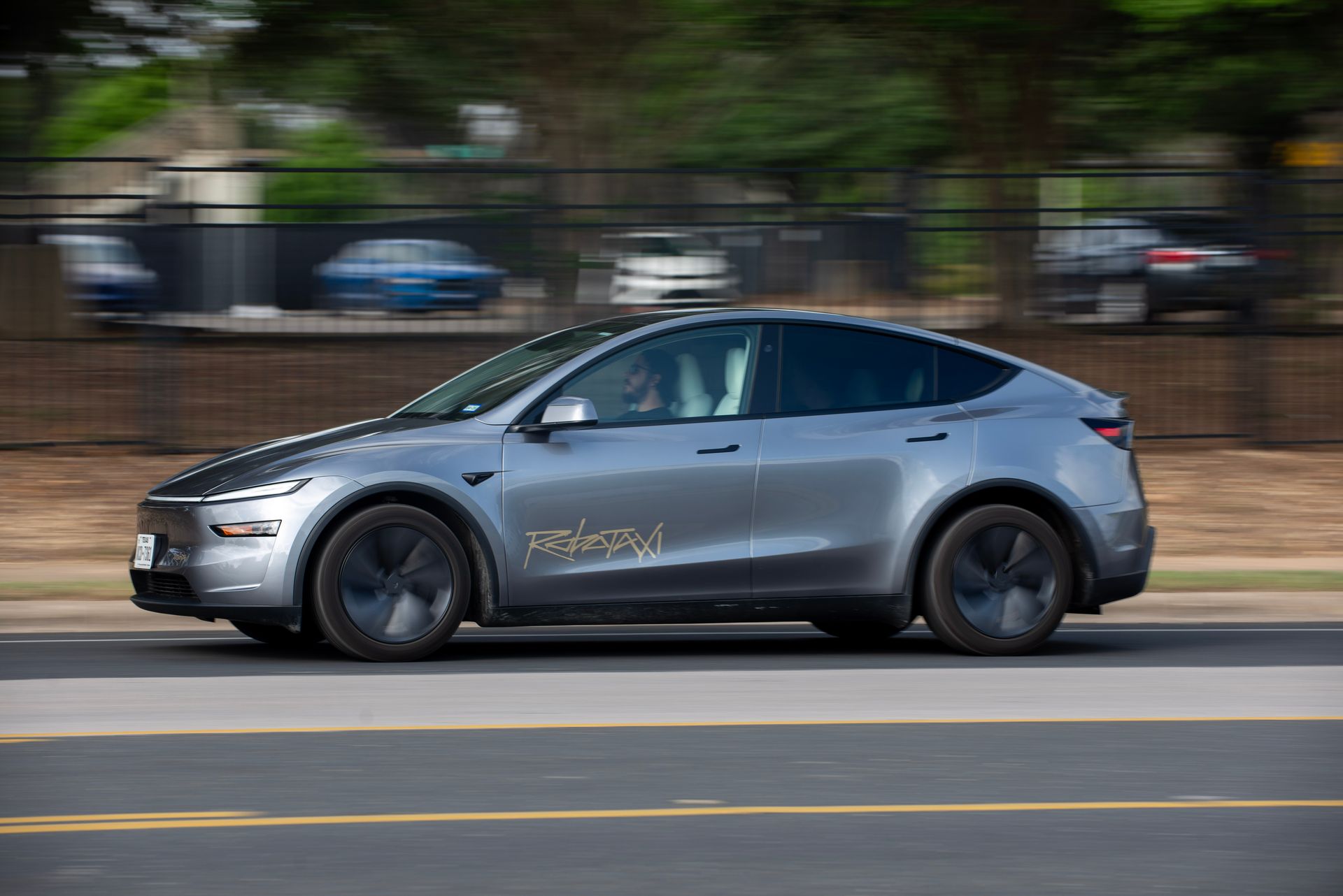 a silver Model Y Tesla SUV with the word "robotaxi" written on it drives down a street