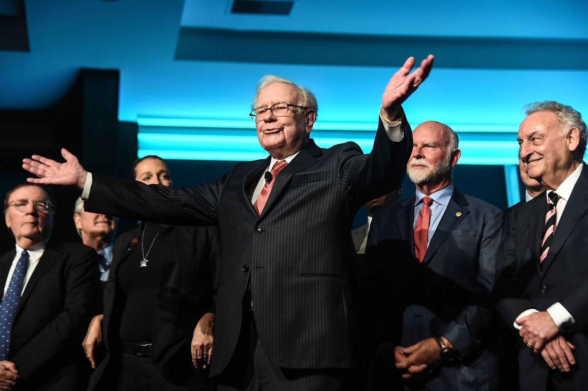 Warren Buffett stands onstage surrounded by a group of people as he lifts and gestures with his arms